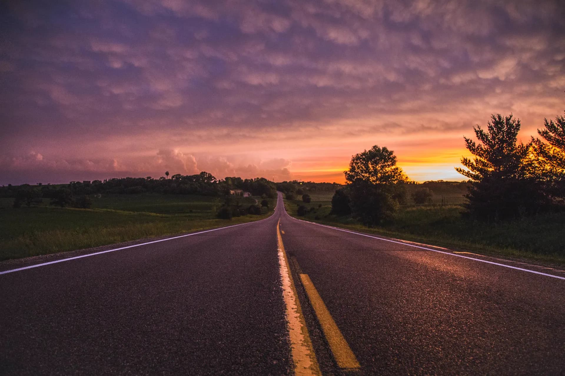 An empty country road running straight through green fields under a golden-hour sky